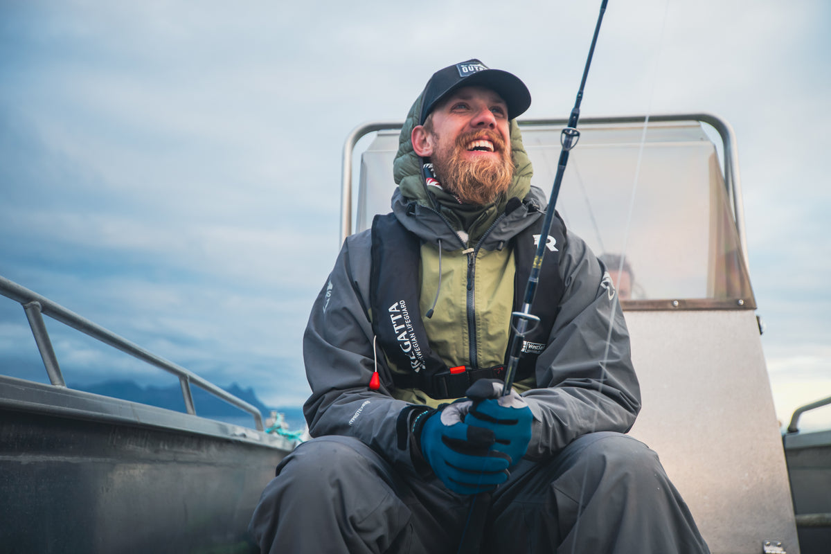 Skäggig man med hatt och handskar sitter leende i båt med fiskespö under fiskeresa, berg och moln i bakgrunden.