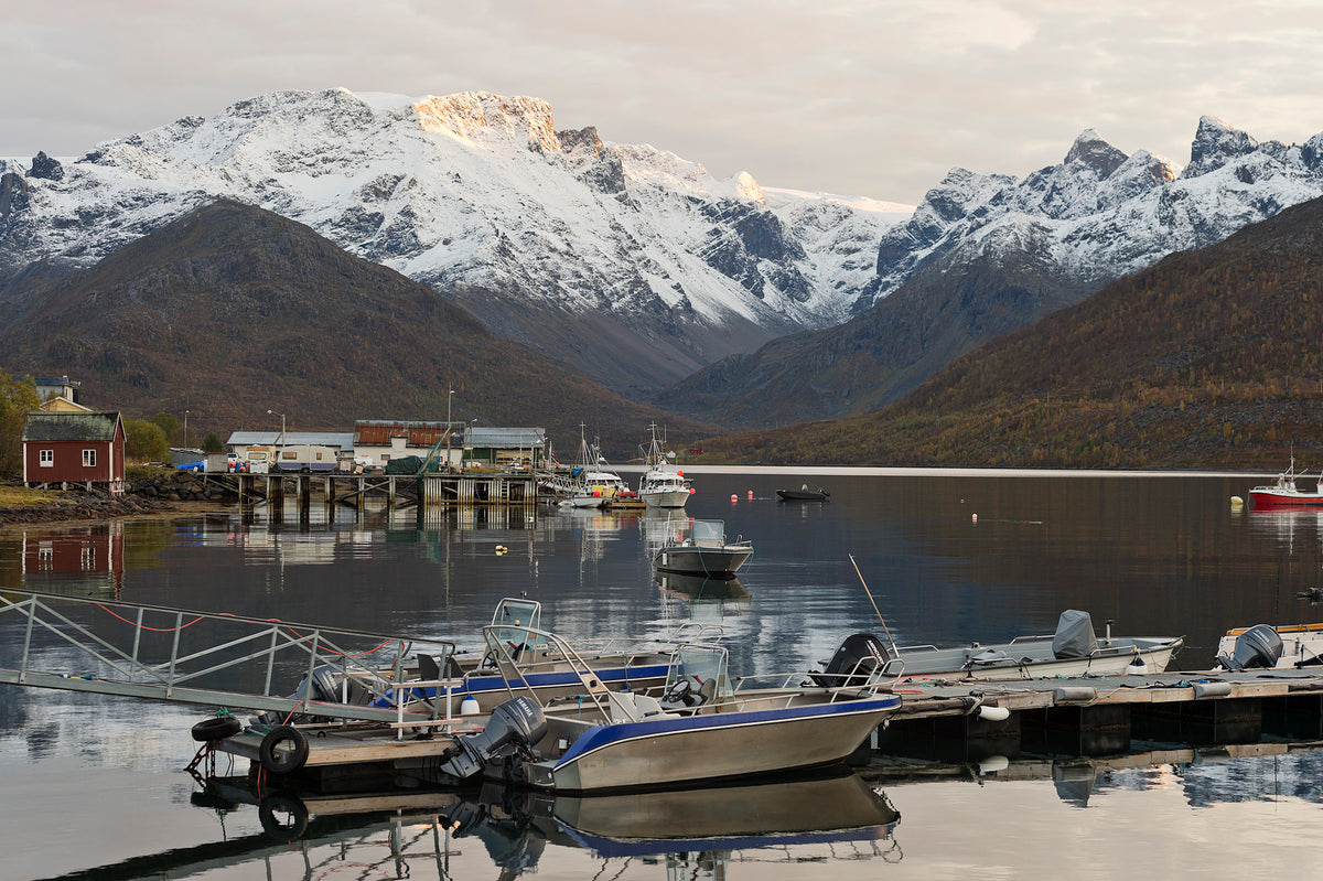 Små båtar vid lugn fjord, snötäckta berg, träbrygga och röda hus - idealiskt för fiskeresa bland höstträd och molnig himmel.