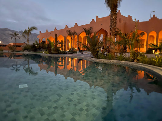 En stilla pool speglar en orange byggnad med valv och palmer i skymningen, berg bakom-perfekt efter sportfiske eller fiskeresa.