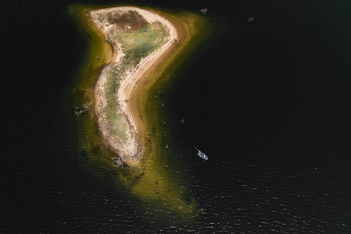 Flygfoto av liten, böjd ö med sandstrand och gräs, omgiven av mörkt vatten. Båt ankrad nära sportfiskare på land och i vattnet.
