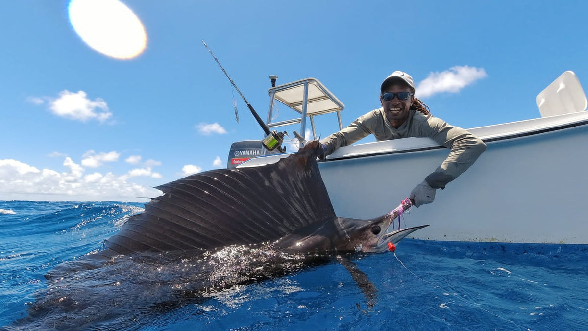 En sportfiskare på båt håller leende en stor segelfisk över havet, ryggfenan syns tydligt mot klar himmel på fiskeresa.