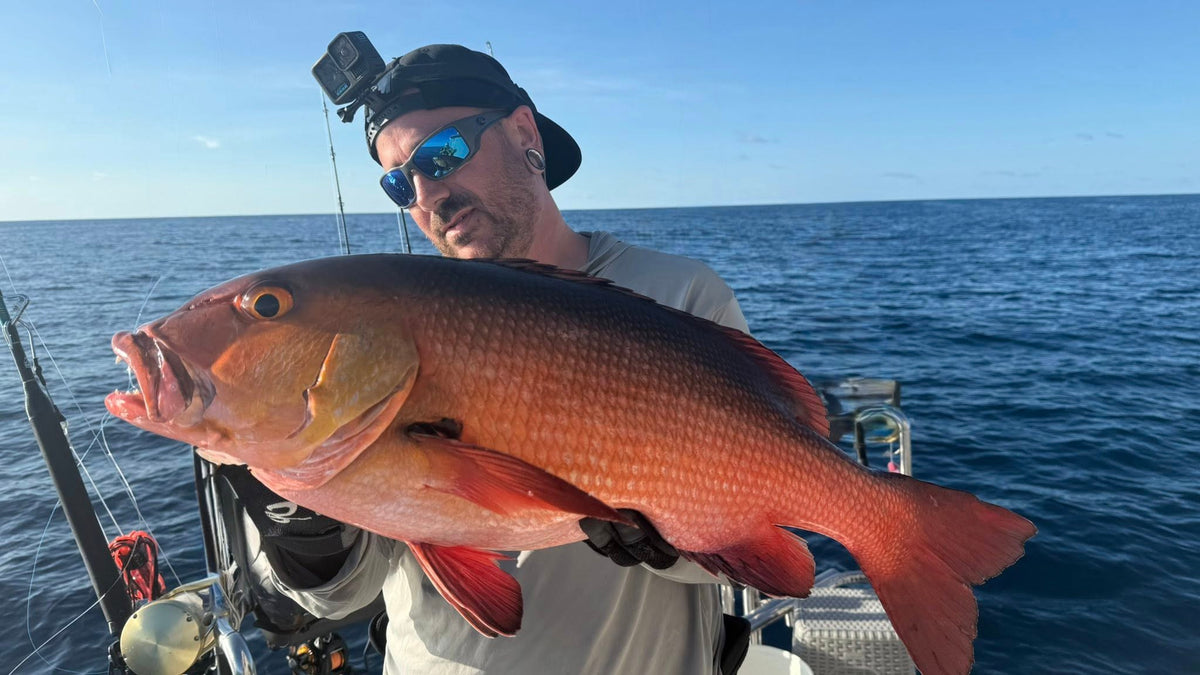 En sportfiskare med solglasögon och bakåtvända kepsar håller upp en stor röd fisk på båt, hav och himmel i bakgrunden.