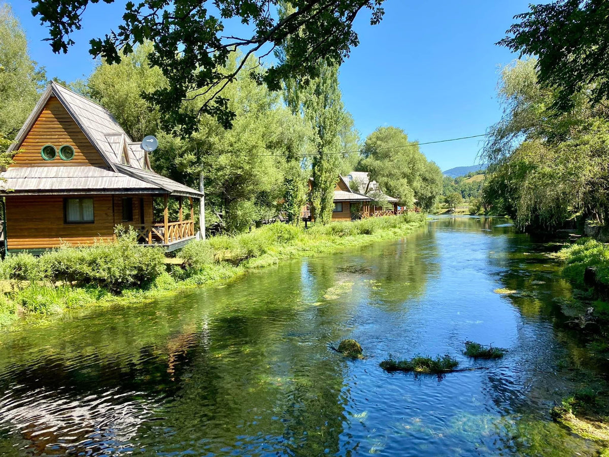 En lugn flod rinner vid trästugor bland gröna träd under blå himmel - idealiskt för fiskeresa och sportfiske i naturen.