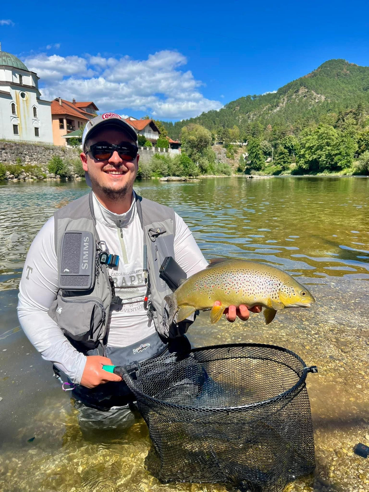 En sportfiskare med solglasögon och fiskväst står i en klar älv, håller en stor öring över håven med fjäll och byggnader i bakgrunden.