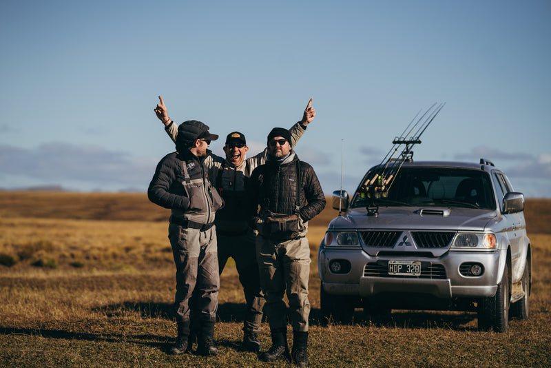 Tre sportfiskare i fiskeutrustning vid en silverfärgad SUV med spön på takräcket, i öppet landskap under klar himmel.
