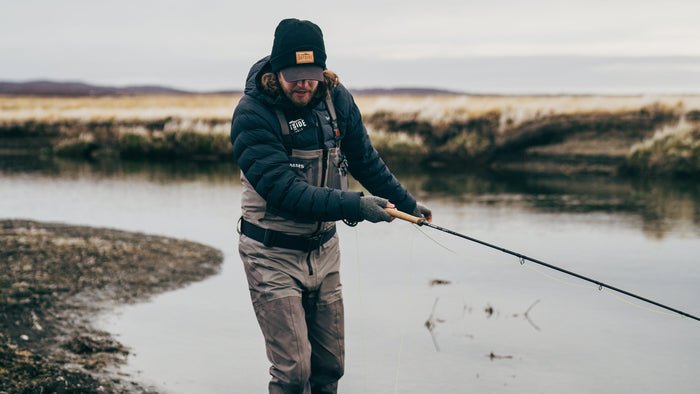 En sportfiskare i mössa, handskar och vadarbyxor fiskar vid en lugn älv. Öppet landskap och molniga skyar i bakgrunden.