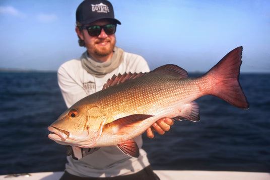 Sportfiskare med solglasögon och keps håller stor röd fisk med vassa fenor på båt under fiskeresa, hav och blå himmel bakom.