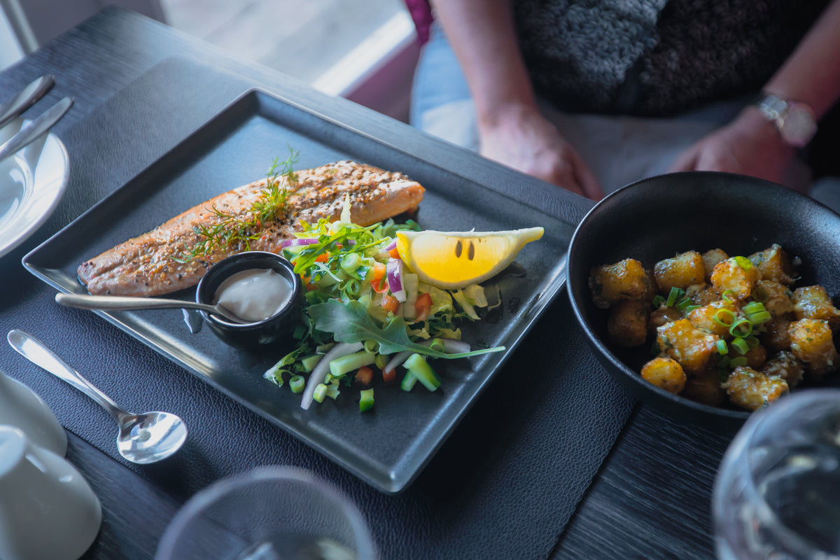 Rektangulär svart tallrik med grillad lax, citron, sallad och sås bredvid skål med rostade potatisar-idealisk efter fiskeresa.