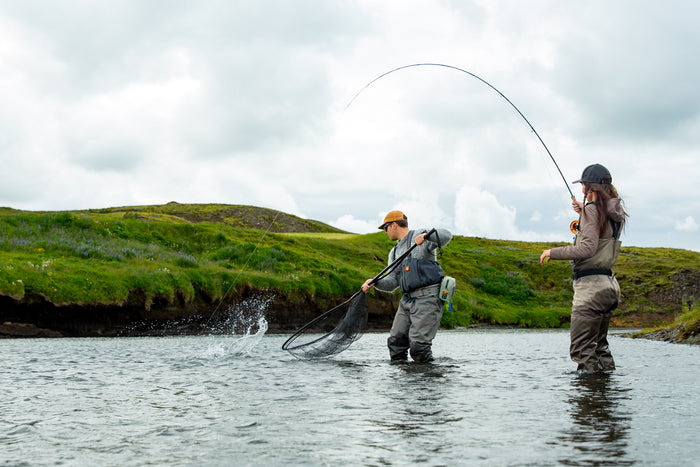 Två personer står i en flod klädda i vadarbyxor; den ena fiskar med ett spö medan den andra hjälper till med håven under deras fisketur bland gröna kullar och porlande vatten.