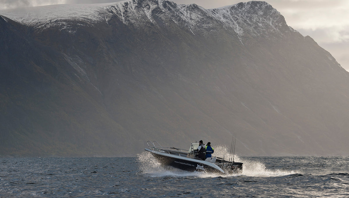 En liten motorbåt med två personer på fiskeresa kör snabbt över vågorna, mot snötäckta berg under molniga himlar.