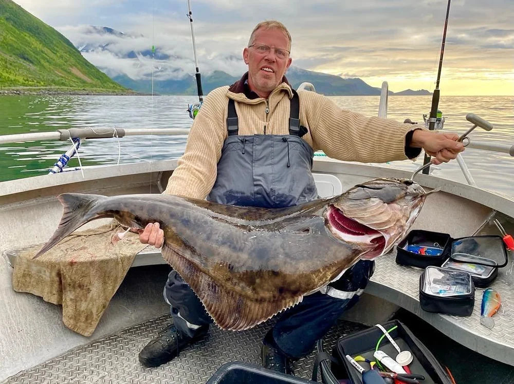 En sportfiskare på båt håller en stor fisk, omgiven av fiskeutrustning och beteslådor med berg och vatten i bakgrunden.