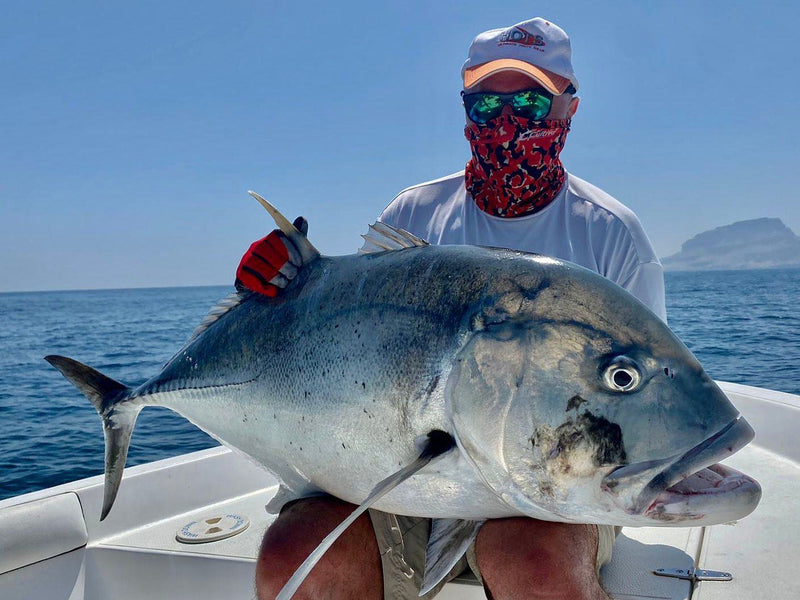 En sportfiskare med hatt, solglasögon och röd ansiktsmask håller en stor fisk i båt på havet med en ö i bakgrunden.