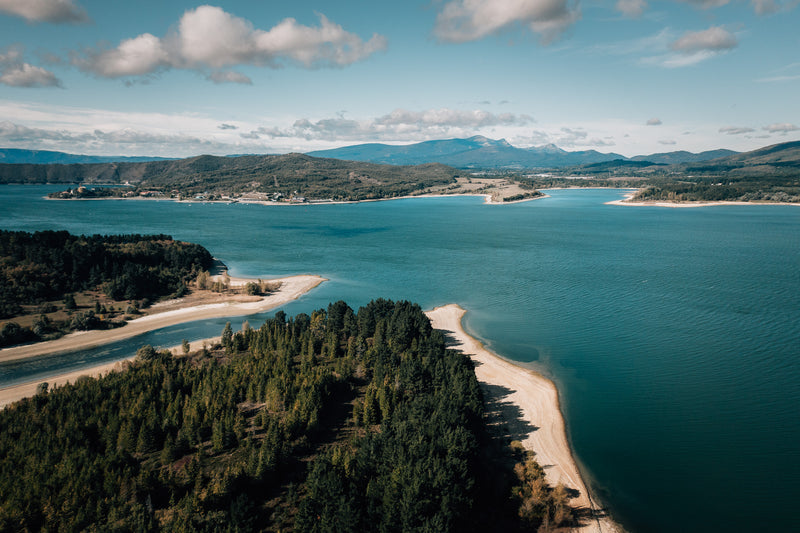 Flygbild över blå sjö med sandstränder och skogsklädda uddar, perfekt för fiskeresa, omgiven av kullar och berg.