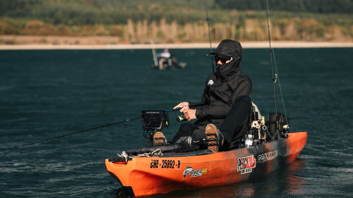 En sportfiskare i orange fiskekajak på sjö, spö i hand, följer ekolod. Ytterligare kajak vid strand under spännande fiskeresa.
