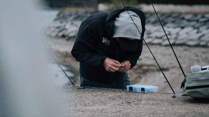 En sportfiskare i huva och keps förbereder fiskeutrustning vid vattnet, med beteslåda och fiskespön under sin fiskeresa.