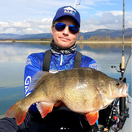 Sportfiskare med solglasögon och blå fisketröja håller stor abborrfisk vid sjö på Fiskeresa Perchzilla i Spanien.