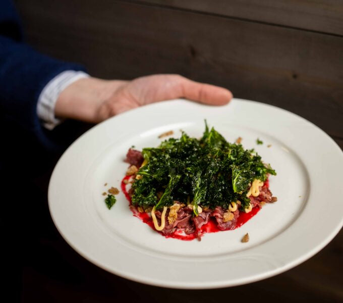 En sportfiskare i blå jacka visar en vit tallrik med tunt skuret kött, krispig grönkål, gul sås och rödbetspuré efter fiskeresa.