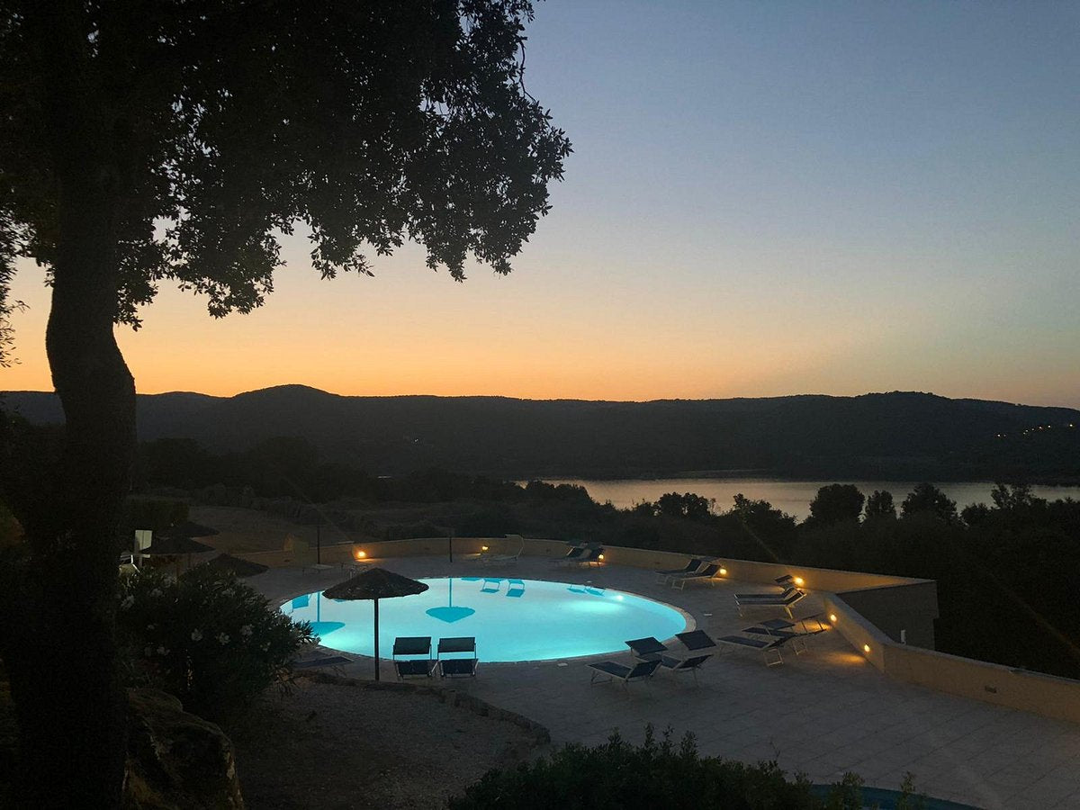 Upplyst pool med solstolar i skymningen, lugn fiskesjö och siluett av kullar. Träd ramar in scenen till vänster.