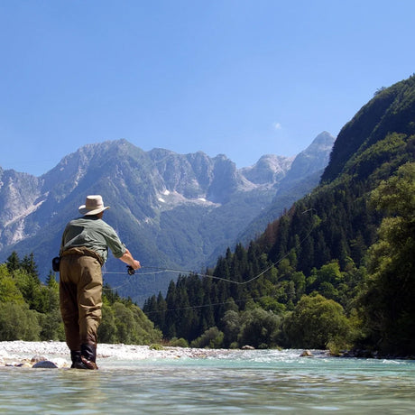 Sportfiskare på fiskeresa i Slovenien flugfiskar i klar älv, omgiven av skog och berg under blå himmel.