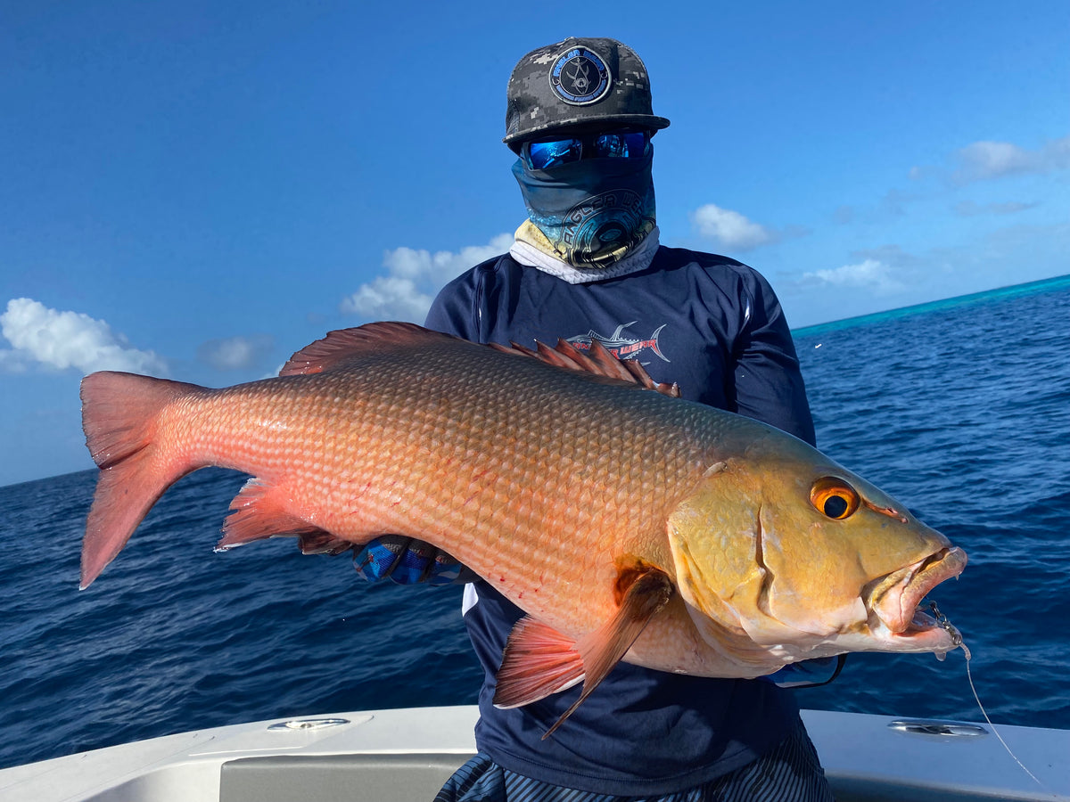 Sportfiskare med hatt, solglasögon och ansiktsskydd håller stor röd snapper på båt, havet och blå himmel i bakgrunden.
