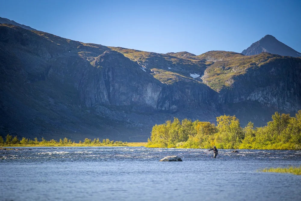 En sportfiskare fiskar i en lugn flod omgiven av gröna träd, höga berg och klarblå himmel - perfekt för fiskeäventyr.
