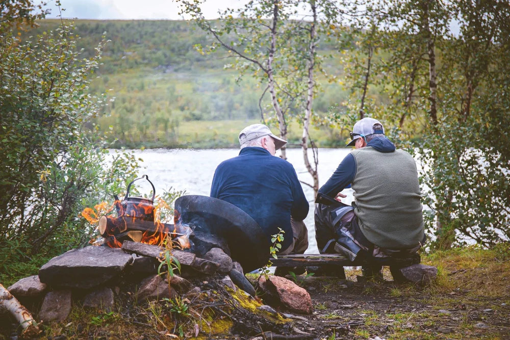 Två personer vid en skogskantad älv värmer vatten över lägereld på fiskeresa, njuter av naturen och gemenskapen.