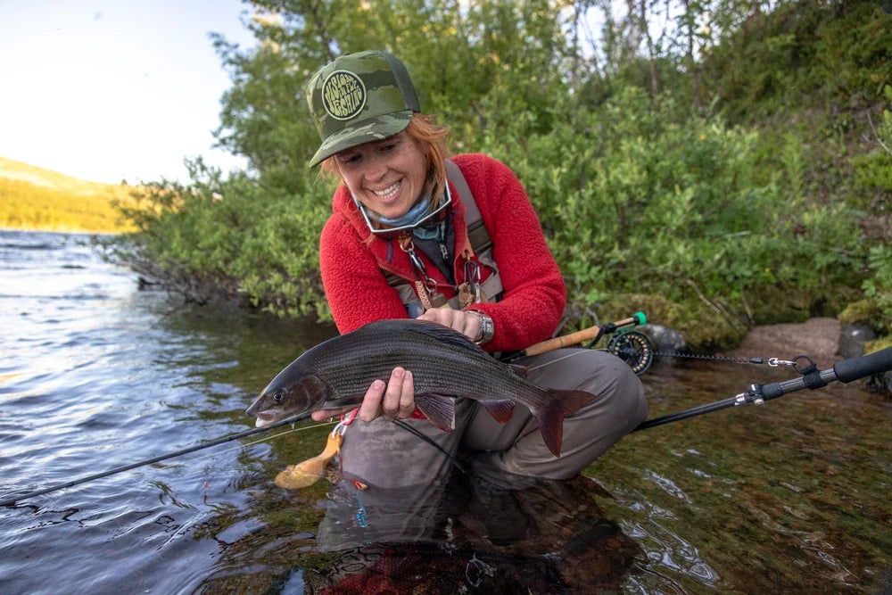 Leende sportfiskare med grön keps vid en älv, håller nyfångad fisk bland fiskeutrustning och grönska under sin fiskeresa.