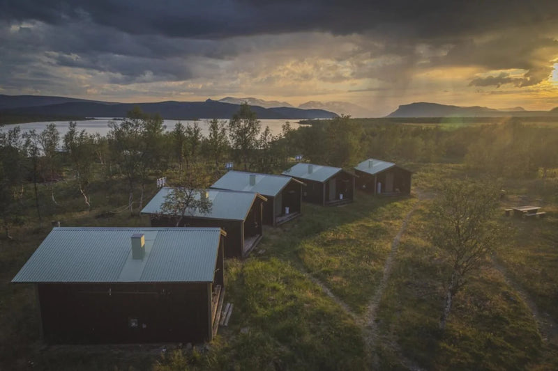 Flygfoto över fem små stugor vid sjö, omgivna av skog och gräs, idealiskt för fiskeresa med fjäll och solnedgång i bakgrunden.