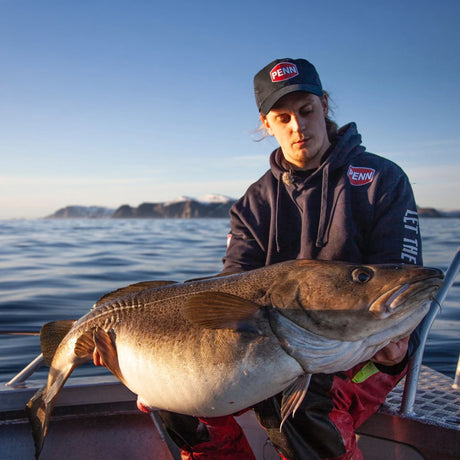 Sportfiskare på båt visar upp stor fisk med hav och snötäckta fjäll i Norge-fiskeäventyr vid Camp Halibut.