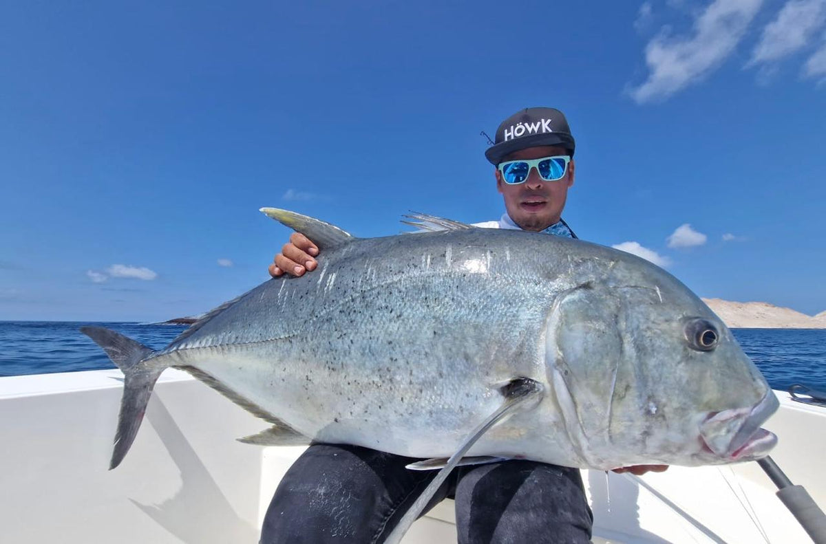 Sportfiskare med solglasögon och keps håller stor fisk på vit båt, omgiven av blått hav och klar himmel under fiskeresan.