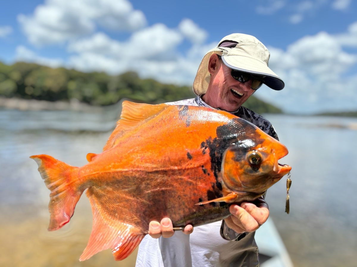 En person med solhatt och solglasögon håller stolt upp en stor, orange fisk vid vattnet, omgiven av träd och moln – en inspirerande bild av en lyckad fisketur.