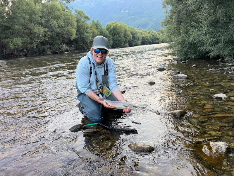 Glad flugfiskare knäböjer i älven med en harr fångad på fluga i Montenegro