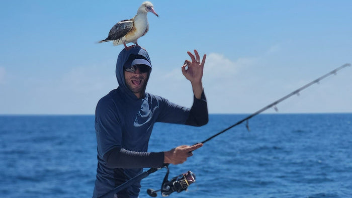 Sportfiskare med solglasögon och blå hoodie håller ett fiskespö på båt, gör OK-tecken, sjöfågel på huvudet, hav och himmel bakom.